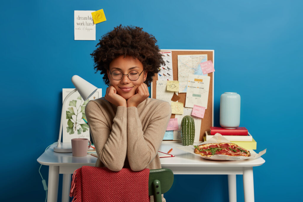 Pretty dark skinned curly woman sits on chair against coworking space, imagines something pleasant, hopes to finish work in time, holds chin and closes eyes.